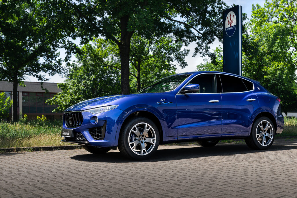 Autofotografie vom exklusiven SUV Maserati Levante in der Farbe Blu Emozione, der auf dem Hof des Autohauses vor Bäumen steht.